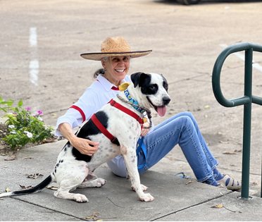 A GJAS volunteer in a straw hat sitting on the sidewalk, hugging a happy black-and-white shelter dog